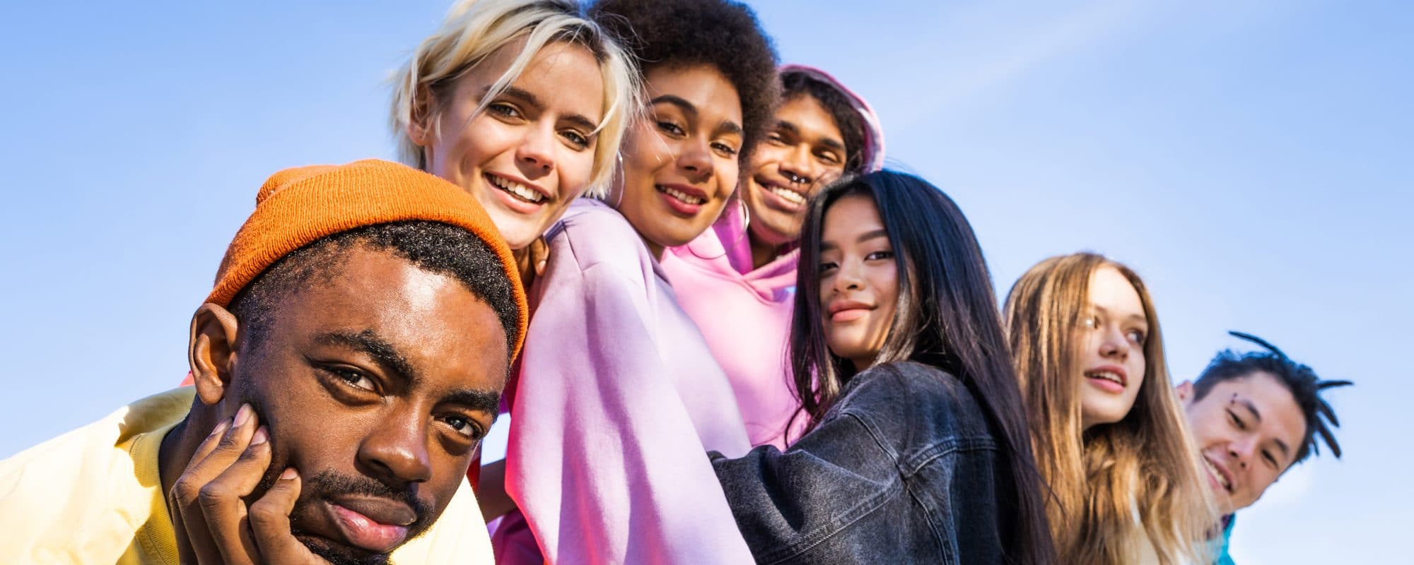 A row of friends from different backgrounds leaning together and smiling under a clear blue sky