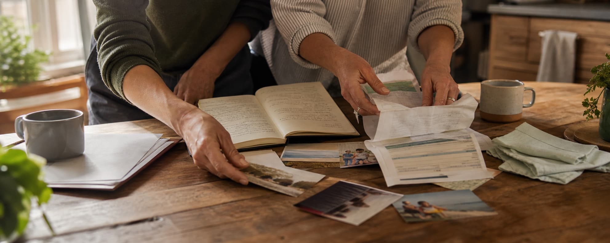Two people sort household papers, notes, and photographs across a sunlit kitchen table